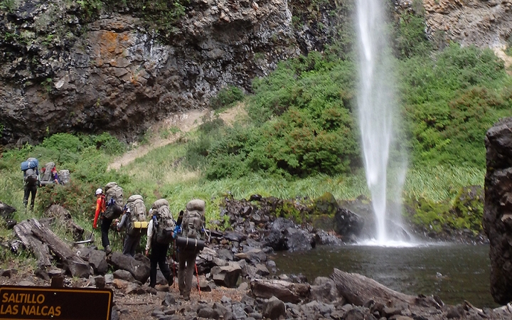 A group of people wearing backpacks hike away from the camera towards a waterfall.